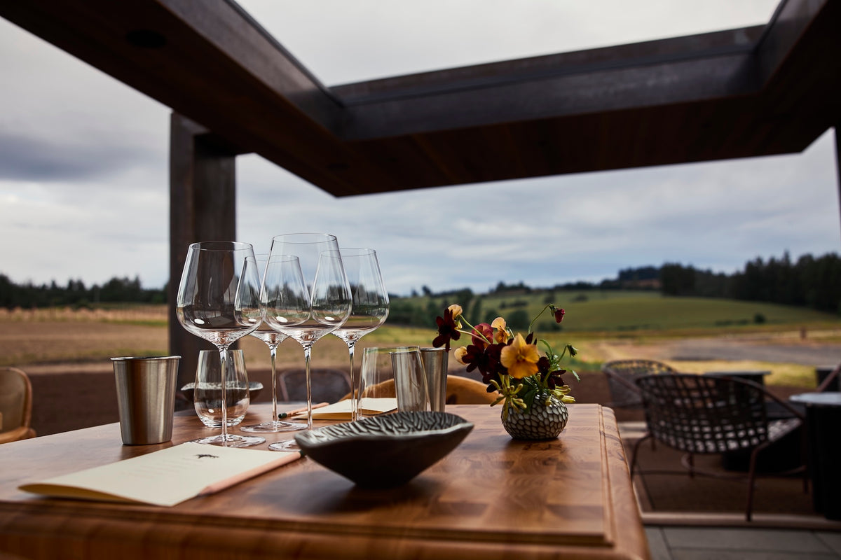 Table with wine glasses and a flower arrangement on a patio with a scenic view.