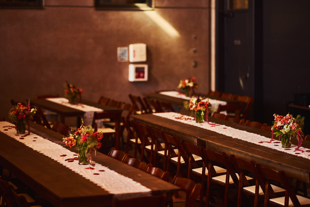 A warmly lit event space with long wooden tables set with white lace table runners, scattered rose petals, and small floral centerpieces, surrounded by wooden chairs against a brown wall.