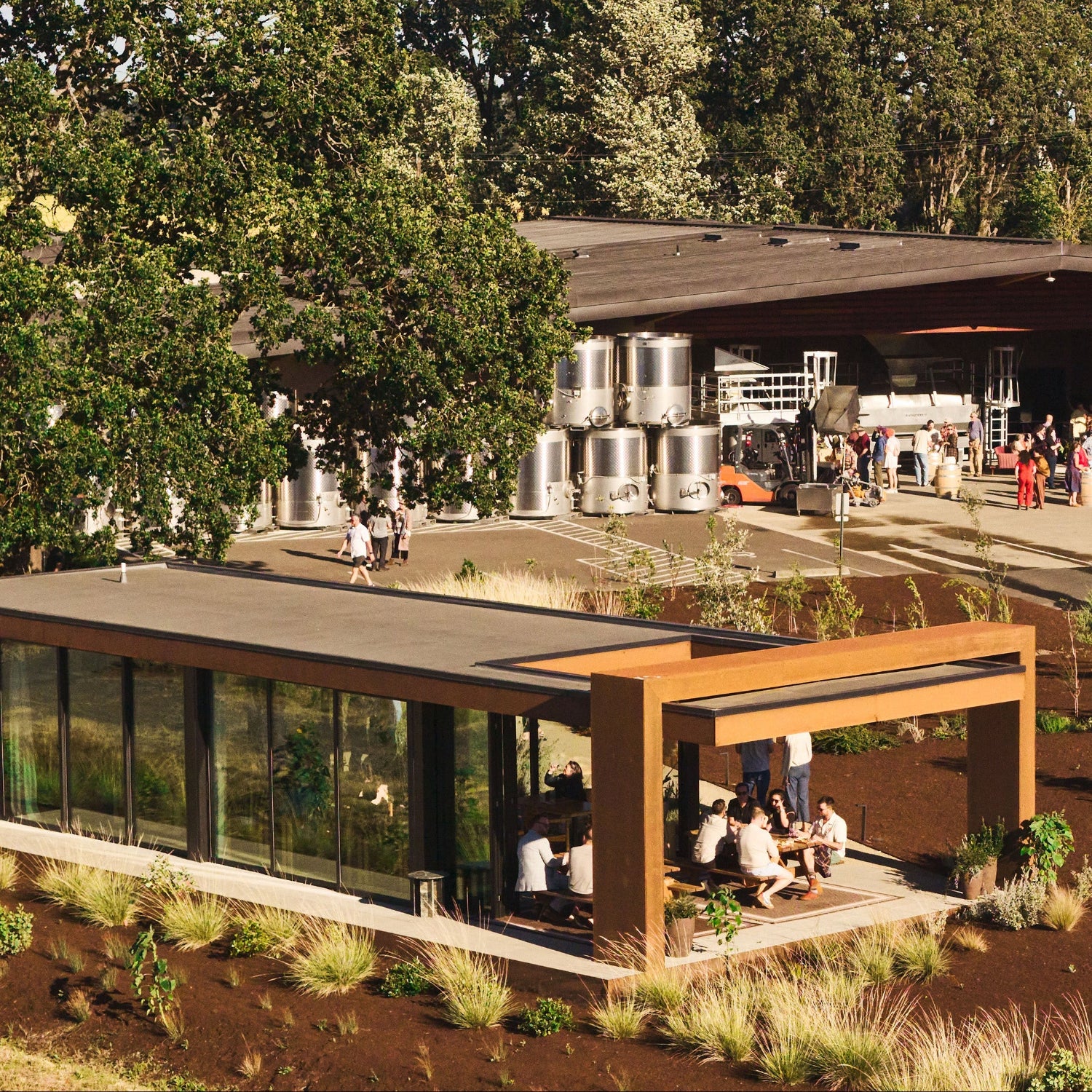 An aerial outdoor scene showing a modern building with a covered seating area in the foreground, a large open workshop or brewery-like structure with stainless steel tanks and many people in the background, and a white arched structure on the right, all surrounded by trees and landscaped ground.
