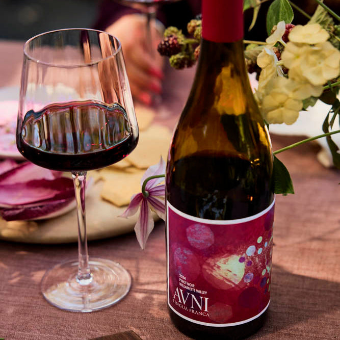 A bottle of Avni red wine with a dark red label, standing on a wooden table next to a filled wine glass, with food and flowers in a styled setting.