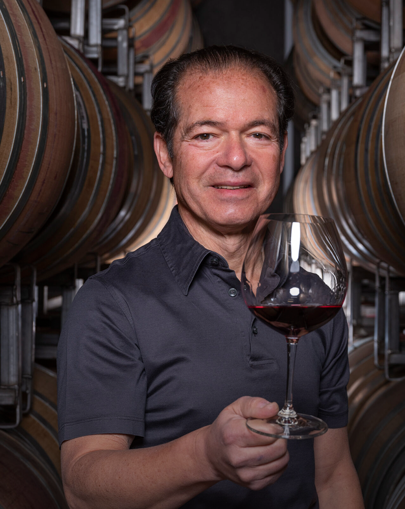 Larry Stone holding a glass of red wine in a winery setting with wine barrels in the background