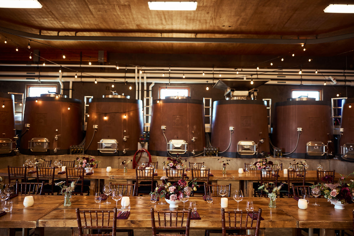 A rustic indoor event space with five large copper-brown distillation tanks in the background, long wooden tables, dark wood chairs, and floral centerpieces with candles set up in a brewery-like room.