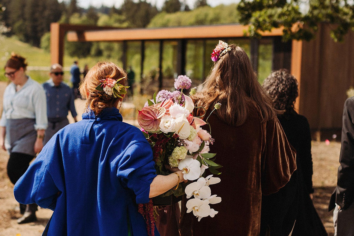 Two women walking outdoors under a tree, viewed from behind. One wears a bright blue top and green skirt while holding a large bouquet of pink, white, and purple flowers.