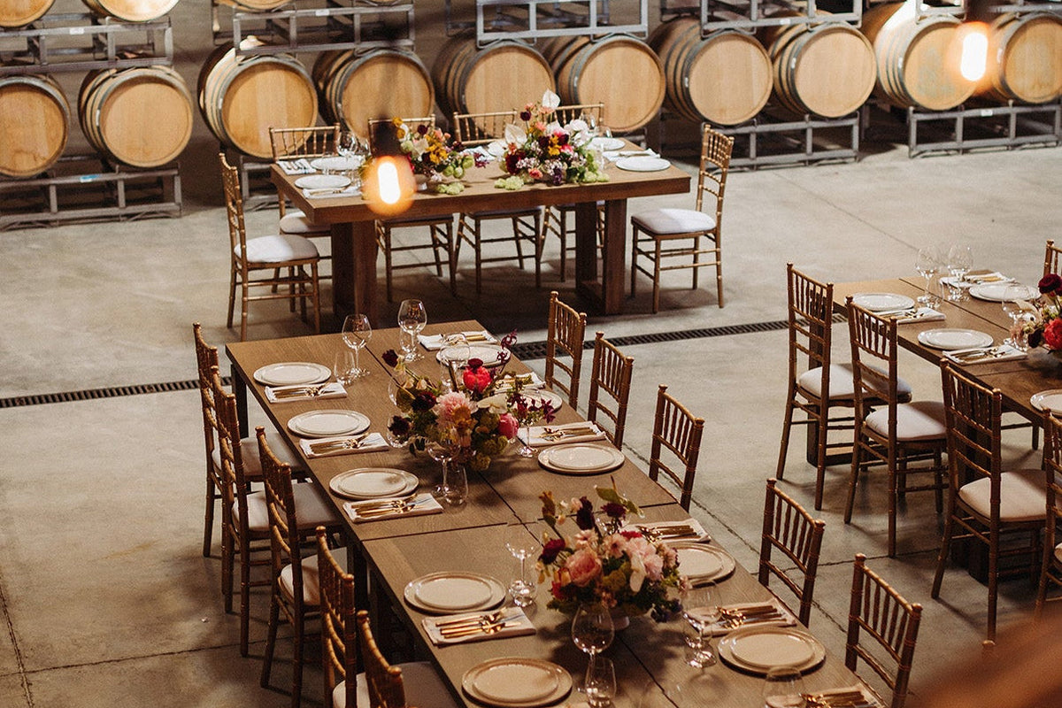 A rustic indoor event space in a wine or brewery setting, with long tables set for dining, wooden chairs, floral centerpieces, and rows of large wooden barrels in the background.